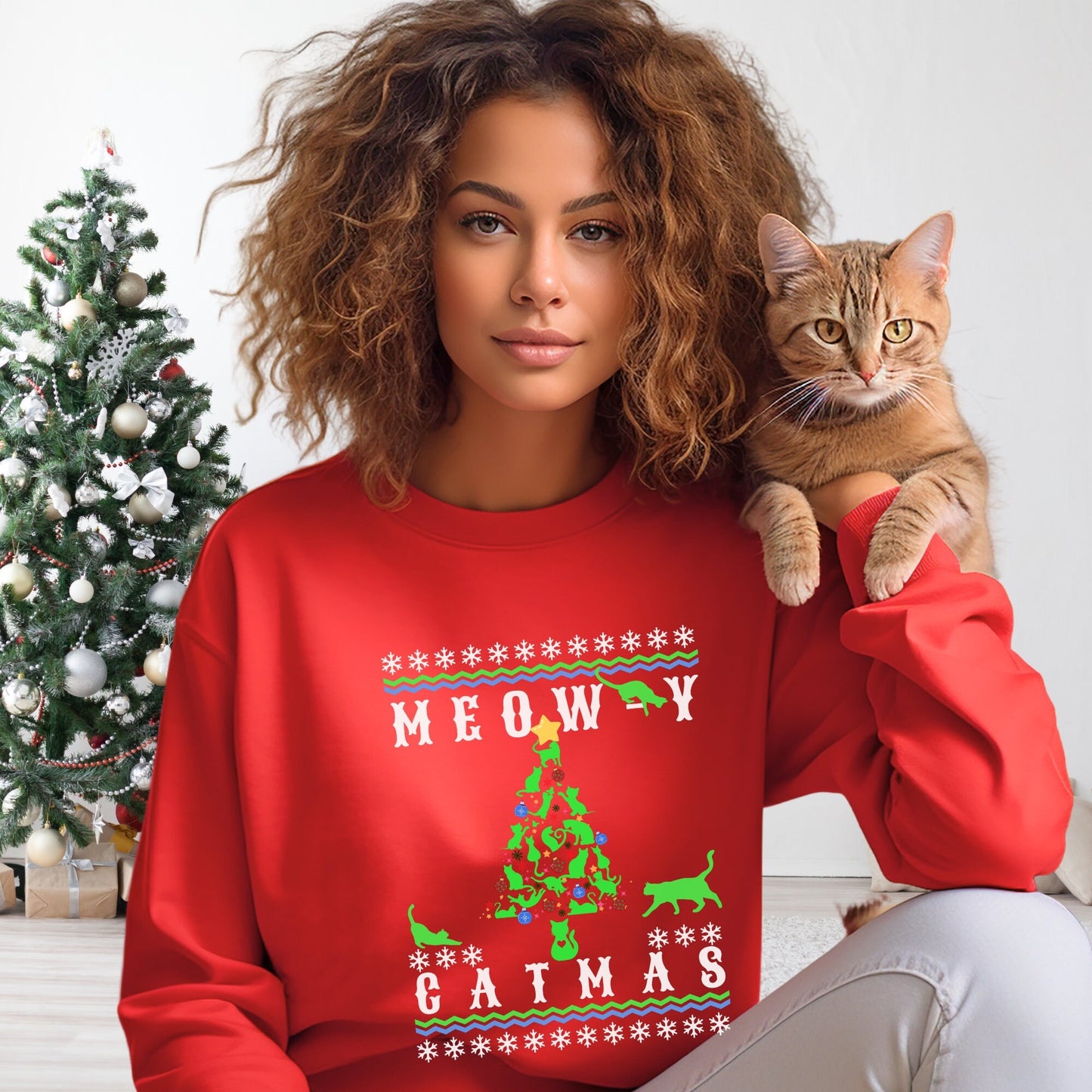 "Model wearing a red 'Meowy Catmas' holiday sweatshirt with a whimsical cat-themed Christmas tree design, posing next to a decorated pine tree."