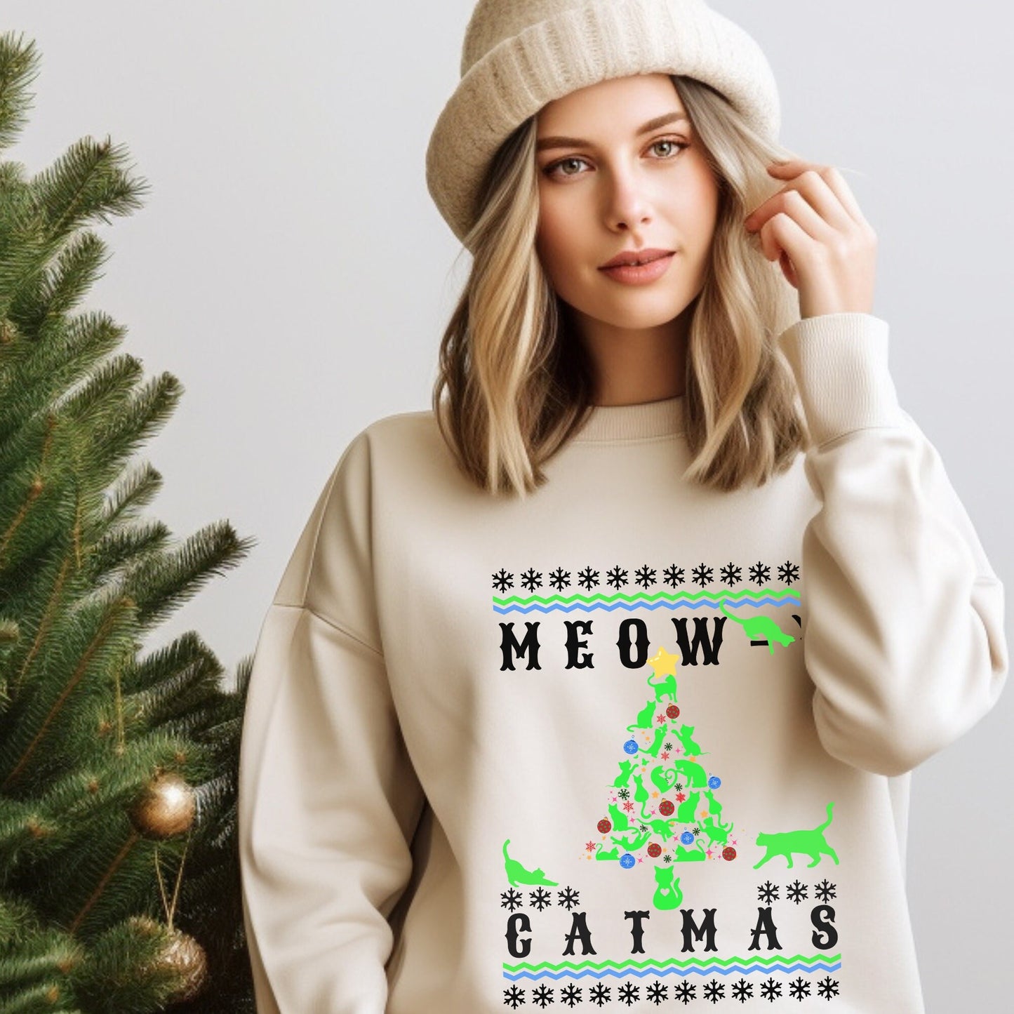 "Model wearing a cream-colored 'Meowy Catmas' holiday sweatshirt with a whimsical cat-themed Christmas tree design, paired with a beige knitted beanie, posing next to a decorated pine tree."
