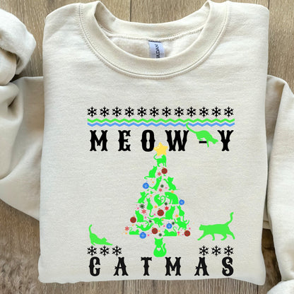 cream-colored &#39;Meowy Catmas&#39; holiday sweatshirt with a whimsical cat-themed Christmas tree design, paired with a beige knitted beanie, posing next to a decorated pine tree.&quot;